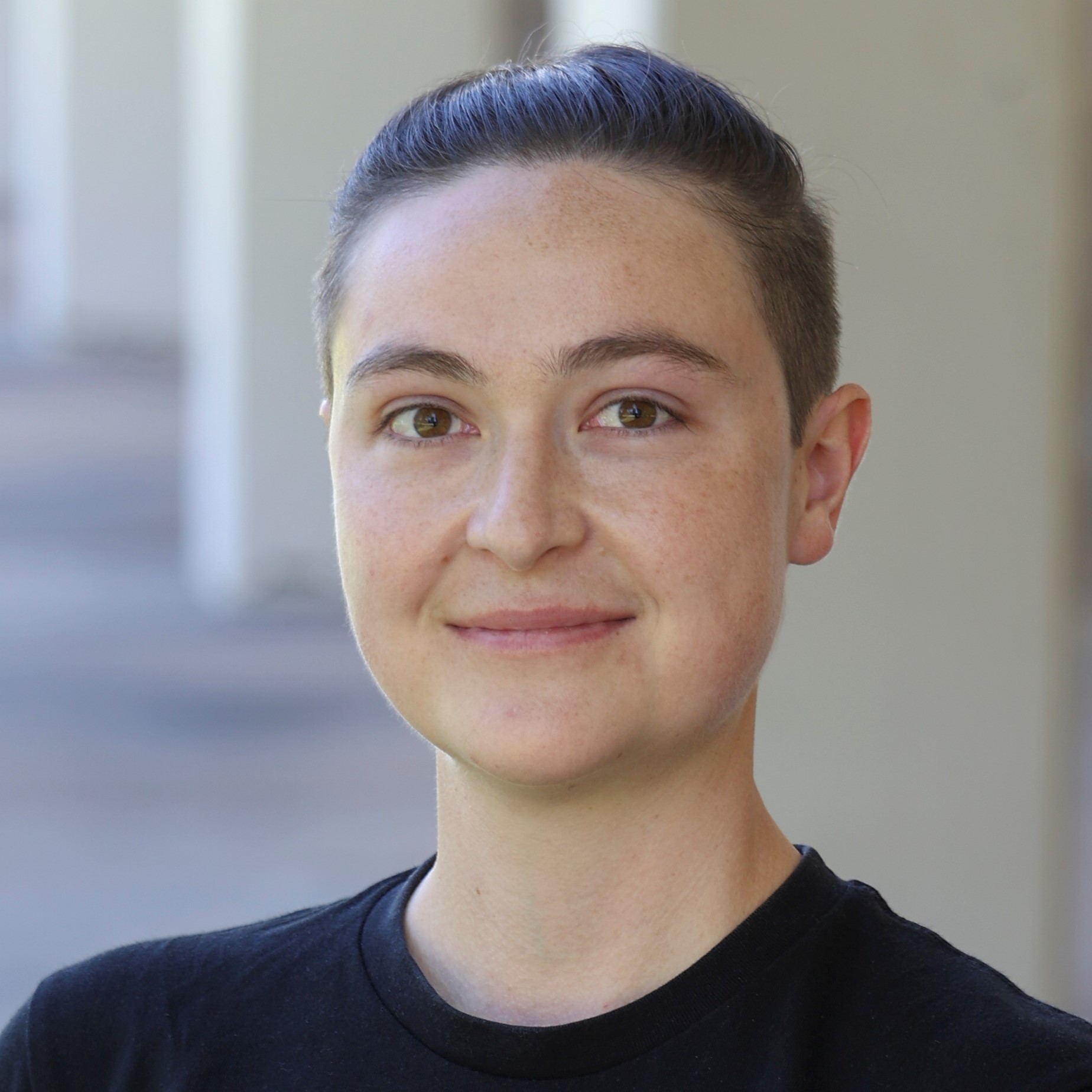 A white person smirks at the camera. They are wearing a black t-shirt and their hair is pulled back in an undercut ponytail.